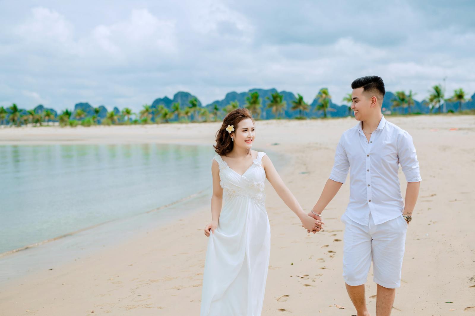 A loving couple walks hand in hand on a tropical beach under cloudy skies.